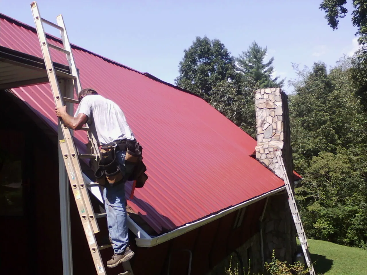 Roofer on a ladder beside a red metal roof and stone chimney — Soffit & Fascia Repair by Roofing Brothers in Quincy